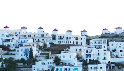 A hillside village composed of whitewashed buildings with blue accents and red-roofed windmills sparsely distributed among the houses. The architecture is traditional and simple, featuring flat roofs and minimalistic design. The overall atmosphere is Mediterranean, likely depicting a scenic island setting.
