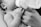A black and white photograph of a small baby being fed from a bottle held by an adult's hand. The focus is on the baby's tiny hand grasping the person's fingers gently.