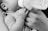 A black and white photograph of a small baby being fed from a bottle held by an adult's hand. The focus is on the baby's tiny hand grasping the person's fingers gently.