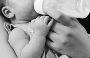 A black and white photograph of a small baby being fed from a bottle held by an adult's hand. The focus is on the baby's tiny hand grasping the person's fingers gently.