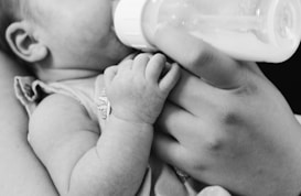 A black and white photograph of a small baby being fed from a bottle held by an adult's hand. The focus is on the baby's tiny hand grasping the person's fingers gently.