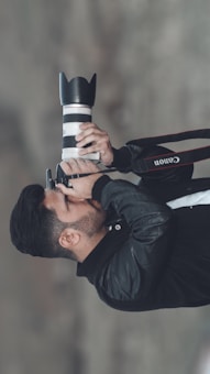A person is holding a camera with a large lens, seemingly capturing a photograph. The person is dressed in a black jacket and is focusing intently through the camera viewfinder.