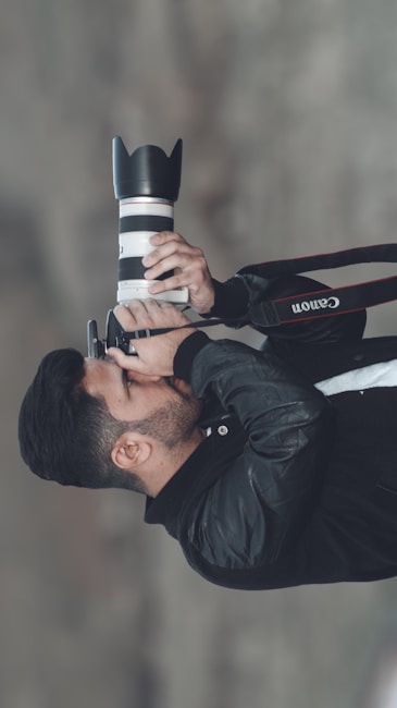 A person is holding a camera with a large lens, seemingly capturing a photograph. The person is dressed in a black jacket and is focusing intently through the camera viewfinder.