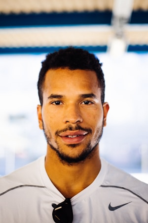 A man with short curly hair and facial hair is wearing a white athletic shirt and has sunglasses hanging from the collar. He is looking directly at the camera with a neutral expression. The background is blurred, suggesting an outdoor or well-lit environment.