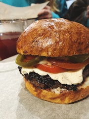 A close-up of a burger with a shiny, golden brown bun. The burger is layered with a thick beef patty, a slice of juicy tomato, melted cheese, and green jalapeño slices. In the background, a glass of dark red beverage is visible along with a blurred figure.