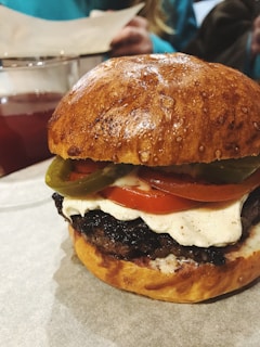 A close-up of a burger with a shiny, golden brown bun. The burger is layered with a thick beef patty, a slice of juicy tomato, melted cheese, and green jalapeño slices. In the background, a glass of dark red beverage is visible along with a blurred figure.
