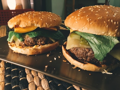 Two delicious-looking hamburgers with sesame seed buns are placed on a tray. Each burger is topped with fresh lettuce, melted cheese, and slices of colorful vegetables. The lighting gives a warm, inviting glow, highlighting the textures of the ingredients. The background features a softly lit room with cozy furnishings.