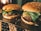 Two delicious-looking hamburgers with sesame seed buns are placed on a tray. Each burger is topped with fresh lettuce, melted cheese, and slices of colorful vegetables. The lighting gives a warm, inviting glow, highlighting the textures of the ingredients. The background features a softly lit room with cozy furnishings.