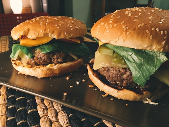 Two delicious-looking hamburgers with sesame seed buns are placed on a tray. Each burger is topped with fresh lettuce, melted cheese, and slices of colorful vegetables. The lighting gives a warm, inviting glow, highlighting the textures of the ingredients. The background features a softly lit room with cozy furnishings.
