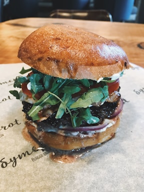 A gourmet hamburger is presented with a glossy bun, fresh arugula leaves, red onion slices, tomato, and a juicy beef patty. The burger is placed on a piece of paper on a wooden table.