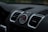 A close-up view of a car dashboard featuring an analog clock with a tachymeter scale in the center. Surrounding the clock are two air vents with a sleek, modern design. The dashboard surface is textured and predominantly black with some metallic accents.