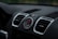 A close-up view of a car dashboard featuring an analog clock with a tachymeter scale in the center. Surrounding the clock are two air vents with a sleek, modern design. The dashboard surface is textured and predominantly black with some metallic accents.