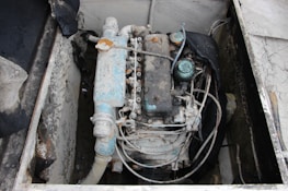 A weathered and rusty engine is situated in a confined space, showcasing various pipes and wires. The metal components exhibit signs of corrosion and wear, with some parts covered in a thin layer of dust or grime. Surrounding the engine is a structure made of what appears to be old concrete or painted metal.