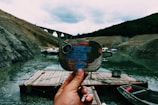 A hand holding an old, rusted can of sardines in front of a serene riverside scene. The background features a wooden raft or dock floating on calm water, several small boats, and a distant arched stone bridge nestled between rugged hillsides under a cloudy sky.