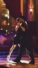 A couple is dancing tango in a dimly lit, elegant room with plush furniture and warm lighting. The woman is wearing a black dress and high heels, and the man is in dark attire. They are in close embrace, focused and graceful.