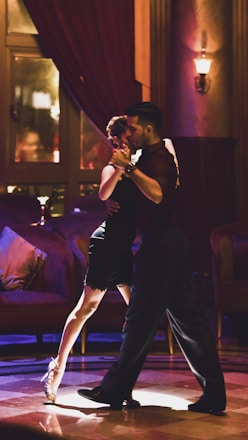 A couple is dancing tango in a dimly lit, elegant room with plush furniture and warm lighting. The woman is wearing a black dress and high heels, and the man is in dark attire. They are in close embrace, focused and graceful.