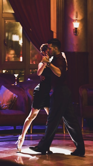A couple is dancing tango in a dimly lit, elegant room with plush furniture and warm lighting. The woman is wearing a black dress and high heels, and the man is in dark attire. They are in close embrace, focused and graceful.