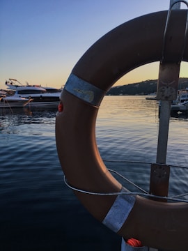 A lifebuoy is prominently displayed in the foreground, attached to a metallic frame. The lifebuoy is brown with worn patches, and a scenic view of a calm body of water is visible in the background. On the water, several boats are anchored, and the horizon showcases a clear sky transitioning from blue to orange, indicating sunset or sunrise.