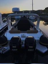 A motorboat is docked at a marina during sunset. The boat has two powerful outboard engines labeled Verado 350. The seating area features brown cushions, and a Garmin device is mounted overhead. Surrounding the boat are other vessels and hills in the background, creating a serene evening atmosphere.