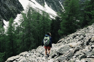 A person with a backpack is hiking on a rocky trail surrounded by pine trees. Snow-covered mountain slopes are visible in the background, highlighting a rugged and natural landscape.