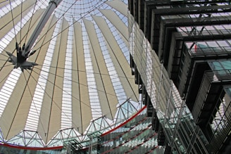 A modern architectural structure featuring a large, circular canopy with radiating fabric panels supported by steel cables. The design appears intricate and futuristic, showing elements of both functionality and aesthetics. In the foreground, there are glass buildings with multiple stories reflecting the intricate patterns of the canopy.