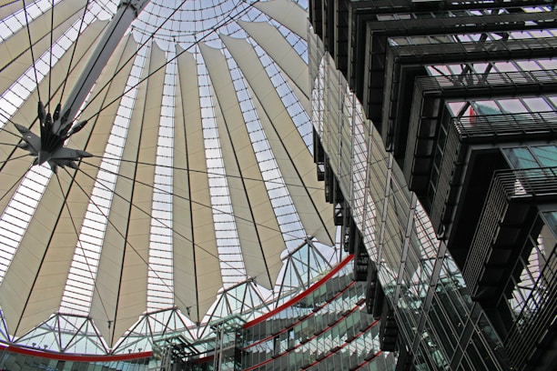 A modern architectural structure featuring a large, circular canopy with radiating fabric panels supported by steel cables. The design appears intricate and futuristic, showing elements of both functionality and aesthetics. In the foreground, there are glass buildings with multiple stories reflecting the intricate patterns of the canopy.