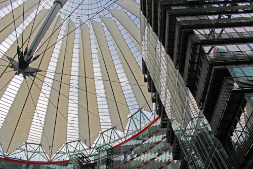 A modern architectural structure featuring a large, circular canopy with radiating fabric panels supported by steel cables. The design appears intricate and futuristic, showing elements of both functionality and aesthetics. In the foreground, there are glass buildings with multiple stories reflecting the intricate patterns of the canopy.