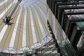 A modern architectural structure featuring a large, circular canopy with radiating fabric panels supported by steel cables. The design appears intricate and futuristic, showing elements of both functionality and aesthetics. In the foreground, there are glass buildings with multiple stories reflecting the intricate patterns of the canopy.