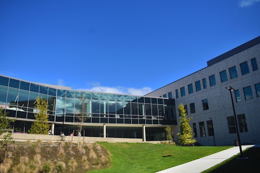 A modern building with large glass windows and a geometric design stands against a clear blue sky. Surrounding the structure is a landscape featuring green grass and young trees. A pathway runs alongside the building, providing access. The architecture includes both glass and solid walls, giving it a sleek and contemporary appearance.