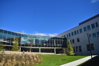 A modern building with large glass windows and a geometric design stands against a clear blue sky. Surrounding the structure is a landscape featuring green grass and young trees. A pathway runs alongside the building, providing access. The architecture includes both glass and solid walls, giving it a sleek and contemporary appearance.