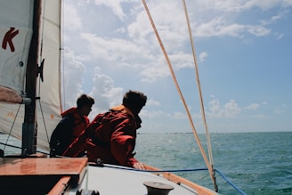 Two individuals wearing life jackets are on a sailboat navigating through a body of water under a partly cloudy sky.