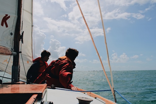 Two individuals wearing life jackets are on a sailboat navigating through a body of water under a partly cloudy sky.