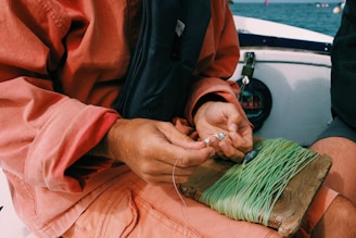 A person wearing a reddish-orange long-sleeve shirt is preparing fishing gear on a boat. Their hands are holding a small metal tool or fishing hook, while a spool of green fishing line is wrapped around a wooden board placed on their lap. The background includes part of the boat and the blue water.