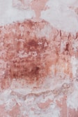 A textured surface with a combination of peeling paint and exposed plaster. The wall features a blend of soft pink and earthy brown tones, creating an aged and weathered appearance.