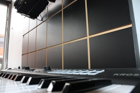 A close-up view of an electronic keyboard or synthesizer, focusing on the keys and control knobs. The background features a wall with a grid pattern, possibly made from acoustic panels. Light enters from a window to the side, creating a bright ambiance.