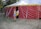 A person is carrying a yellow basket and entering a large red and yellow circus tent from the side. The tent is situated outdoors on a grassy area surrounded by trees. Various ropes and poles are used to secure the tent in place.