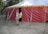 A person is carrying a yellow basket and entering a large red and yellow circus tent from the side. The tent is situated outdoors on a grassy area surrounded by trees. Various ropes and poles are used to secure the tent in place.
