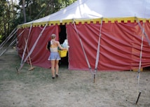 A person is carrying a yellow basket and entering a large red and yellow circus tent from the side. The tent is situated outdoors on a grassy area surrounded by trees. Various ropes and poles are used to secure the tent in place.