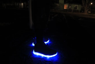 A pair of shoes with bright blue LED lights on the soles glowing in the darkness. The background is dimly lit, suggesting a nighttime setting with faint silhouettes of buildings and trees.