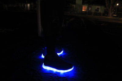 A pair of shoes with bright blue LED lights on the soles glowing in the darkness. The background is dimly lit, suggesting a nighttime setting with faint silhouettes of buildings and trees.