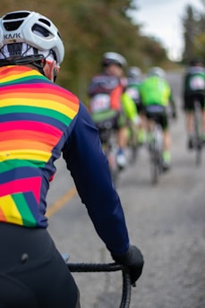 A group of cyclists riding on a road, with the focus on a rider wearing a rainbow-colored jersey and a white helmet. Other riders in various colored jerseys are in the background, slightly out of focus.