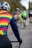 A group of cyclists riding on a road, with the focus on a rider wearing a rainbow-colored jersey and a white helmet. Other riders in various colored jerseys are in the background, slightly out of focus.