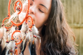 A person with long, wavy hair is holding a colorful dreamcatcher adorned with feathers and beads close to their face. The dreamcatcher features several circular webs and a combination of red, black, and white feathers. The background is slightly blurred, suggesting an outdoor setting.