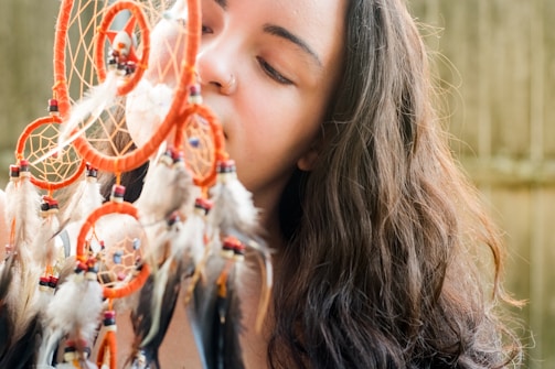 A person with long, wavy hair is holding a colorful dreamcatcher adorned with feathers and beads close to their face. The dreamcatcher features several circular webs and a combination of red, black, and white feathers. The background is slightly blurred, suggesting an outdoor setting.