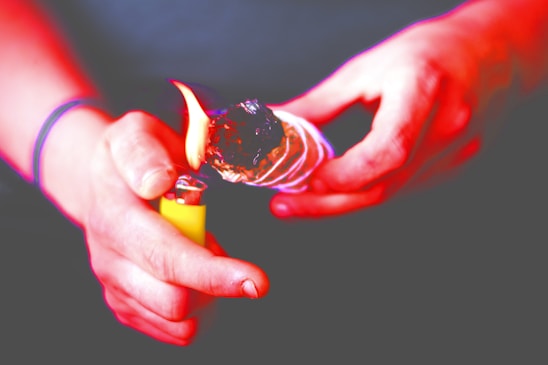 Hands holding a bundle of sage that is being lit by a yellow lighter. The sage appears to be burning, with a visible flame at the tip. The background is out of focus, emphasizing the ritualistic action in the foreground.