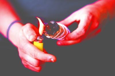 Hands holding a bundle of sage that is being lit by a yellow lighter. The sage appears to be burning, with a visible flame at the tip. The background is out of focus, emphasizing the ritualistic action in the foreground.