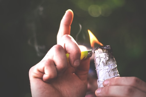 A hand holding a lighter is igniting a bundle of sage wrapped with white string. Smoke is faintly visible as it rises from the burning sage. The background is softly blurred with a dark, natural ambiance suggesting an outdoor setting.