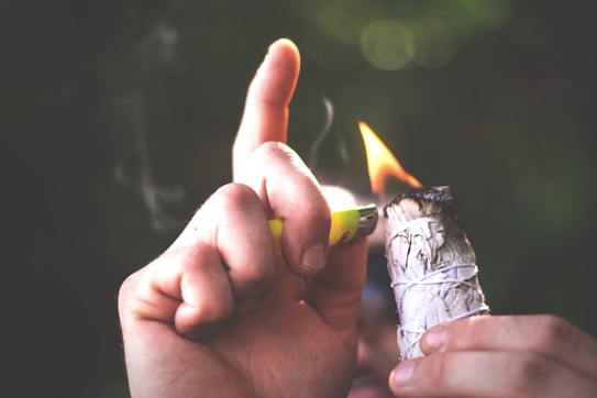 A hand holding a lighter is igniting a bundle of sage wrapped with white string. Smoke is faintly visible as it rises from the burning sage. The background is softly blurred with a dark, natural ambiance suggesting an outdoor setting.