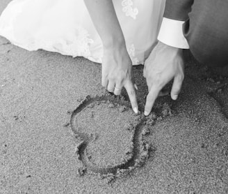 grayscale photography of man and woman draw heart on sand