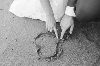 grayscale photography of man and woman draw heart on sand